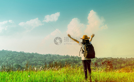 女爬坡道带着帽子和背包站的快乐女游客在山上举起手来在阳光日照耀的子用绿草地举起手来用蓝天和积雪云彩旅行者享受美丽的风景户外图片