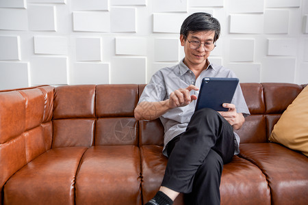 快乐的互联网高级亚裔老人在家中客厅沙发上玩平板和视频电话的亚洲老年人长者肖像正在放松与幸福并用平板游戏图片