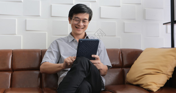 亚裔老人在家中客厅沙发上玩平板和视频电话的亚洲老年人长者肖像正在放松与幸福并用平板游戏电脑人们眼镜图片