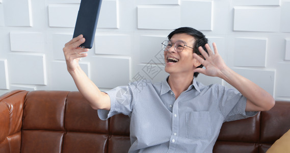 微笑亚裔老人在家中客厅沙发上玩平板和视频电话的亚洲老年人长者肖像正在放松与幸福并用平板游戏闲暇笑图片