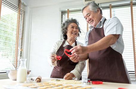 手快乐微笑的老年情人男和女在厨房烘烤迷人的老夫妇享受抱着红饼干切机心脏形状的红饼干切割机在家里一起准备和敲鼓钱乐趣曲奇饼图片