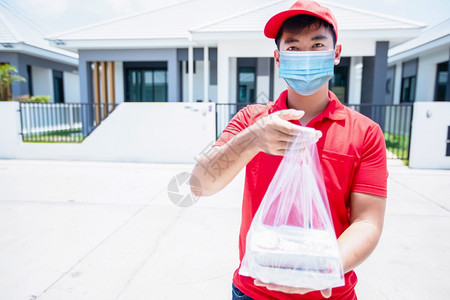 航运男人亚洲送货服务员身穿红色制服戴着帽子的制服并面戴罩用塑料袋处理食品箱在房子门前交给顾客在线购物和快递邮差图片