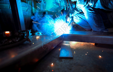 劳动作坊热的焊工用氩弧机接金属并产生火花一名男子戴防护手套工业作场所的安全焊工与人在钢铁厂作图片