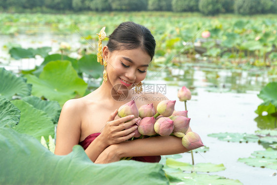 年轻的户外身着传统泰式礼服坐在花莲湖的木船上她手握着粉红莲水捡起花朵她的脸很快乐笑容好声泰国图片
