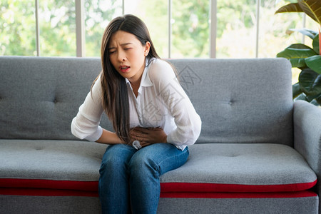 恶心亚洲人下午坐在沙发上并忍受腹部因月经泻或消食不良而痛的腹部疼不快亚裔妇女坐落在沙发上忍受着腹部疼痛疾病和保健概念背景图片