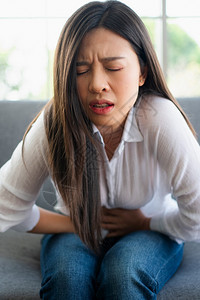 坐在沙发上并忍受腹部因月经泻或消食不良而痛的腹部疼不快亚裔妇女坐落在沙发上忍受着腹部疼痛疾病和保健概念炎恶心康背景图片