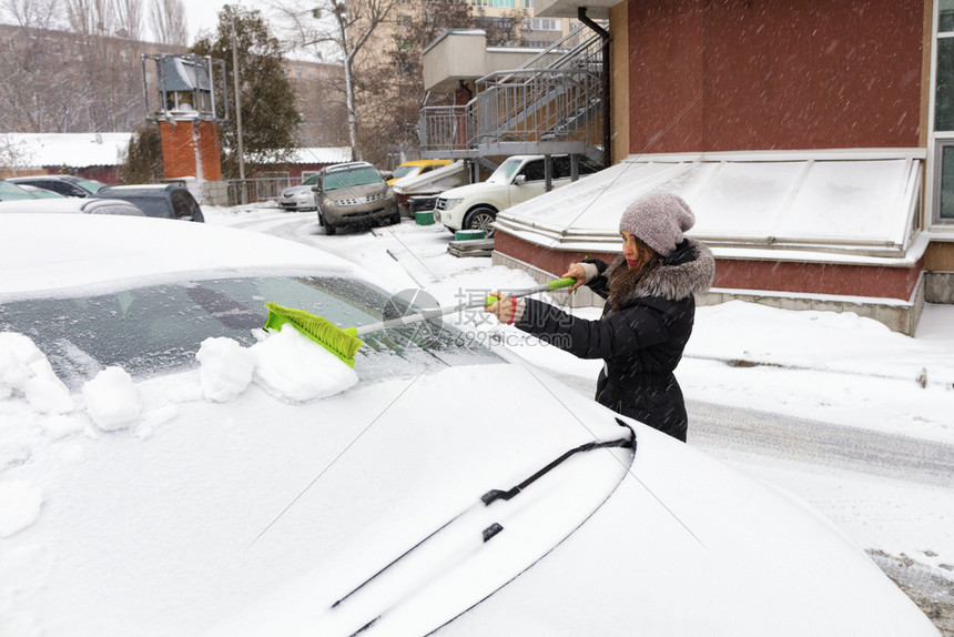 汽车人苍白身穿黑大衣的年轻美女用一辆汽车表面折叠的绿色长梳子扫雪年轻妇女用车表面的绿刷子擦雪年轻妇女用车表面净雪屋图片