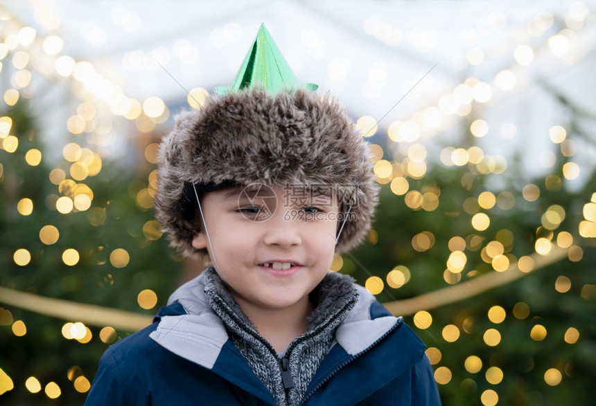 新的十二月快乐可爱男孩带着微笑的脸戴党帽面对圣诞树上模糊的灯光Kid在传统圣诞节市场庆祝活动上玩得开心孩子站在明亮的Xmas树旁图片