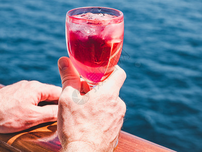 在蓝海浪背景下游轮露天甲板上盛着一杯美丽的粉红葡萄酒时装男子侧观特近休闲和旅行概念暇和旅游概念持有美丽粉红葡萄酒的人日落派对精图片