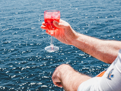 红色的商业在蓝海浪背景下游轮露天甲板上盛着一杯美丽的粉红葡萄酒时装男子侧观特近休闲和旅行概念暇和旅游概念持有美丽粉红葡萄酒的人男图片