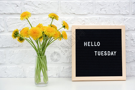 你好星期二在黑色信板上的单词和桌子一束黄色蒲公英花在白砖墙上朵明亮的早晨图片