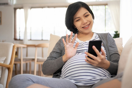 美丽的亚洲孕妇在家中怀时网上进行视频聊天通过智能电话交谈和与丈夫及家人握手快乐笑的年轻母亲用手机享受其中的快乐之情保持产假联系图片