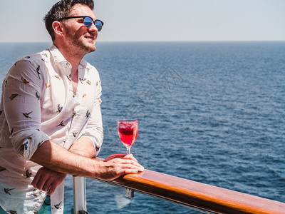 太阳在蓝海浪背景下游轮露天甲板上盛着一杯美丽的粉红葡萄酒时装男子侧观特近休闲和旅行概念暇和旅游概念持有美丽粉红葡萄酒的人一种蓝色图片