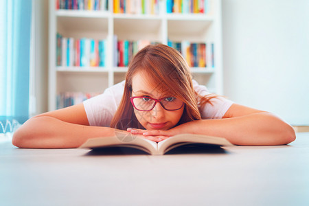 读者相机坐在家庭研究书架前的地板上或头戴眼镜看着用亲手阅读的年轻漂亮女生学一身戴眼镜看头闲暇图片