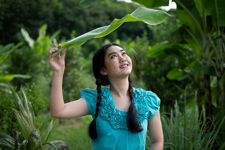 抓住快乐的衬衫一位亚洲年轻女肖像身着黑头发在绿色花园背景上拿着香蕉叶图片