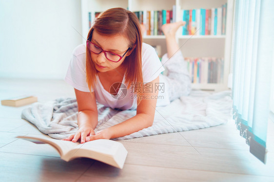 快乐的在书架前躺的地板上身戴眼镜持有一本书的年轻美女学生在家里阅读或学习的肖像教育20多岁图片