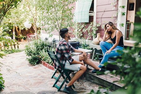 女朋友娱乐肖像用智能手机坐在木屋附近的花园椅子上微笑的年轻夫妇他们用智能手机在他们的木屋附近坐着花园椅图片