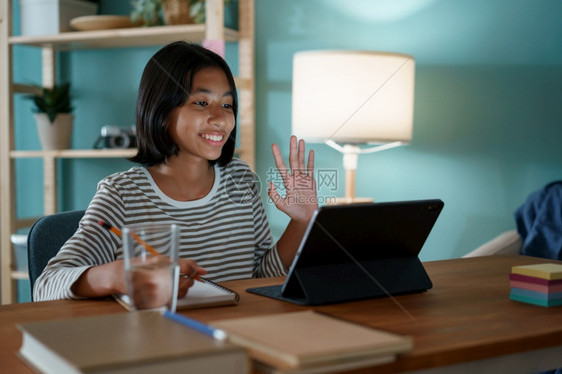 班级快乐的亚洲女孩通过互联网在上学习使用平板手挥舞贺礼的问候牌子在家晚上坐办公桌边亚洲儿童观察做家庭作业女夜晚图片