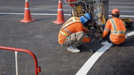 道路指示标热塑料金属在职的两名穿反射背心的公路工人正在修补部分热塑料喷雾式道路标识机器上面装有交通锥形和沥青路红屏障用于铺设交通线油漆工作背景