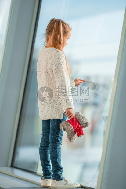 离开游客在大窗户附近的机场带着泰迪熊玩具的小女孩寻找大飞机在窗户附近的机场等待登的小女孩在等着登机人们图片