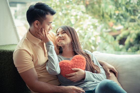 年轻的女婚姻在沙发上坐着的亚洲情侣们妇女怀着红色的心和欢笑快乐微爱表达和人温暖概念情人节图片