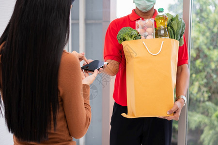 一种亚洲送货员持有一袋新鲜食品供顾客使用并拥有智能手机在家中接收付款快速杂货服务和新生活方式概念a提供快餐服务和新生活方式的概念图片