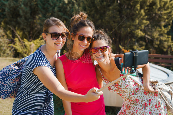 摄影幸福三名年轻女游客使用自拍棍和手机智能在阳光明媚的秋天穿着暑期服装进行自我肖像晴天图片