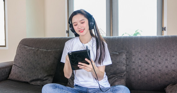 白色的手机请享用开心快乐的亚洲年轻女子可爱的美丽戴着耳机从平板听音乐她正在玩平板和坐沙发上家里笑声图片