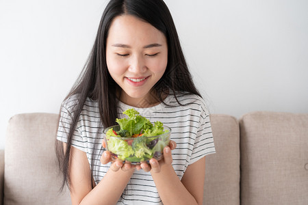 吃亚洲妇女坐在沙发上享受健康和生活方式概念时却坐在家里的沙滩上手持一碗盛满绿色沙拉蔬菜的玻璃碗微笑年轻的图片