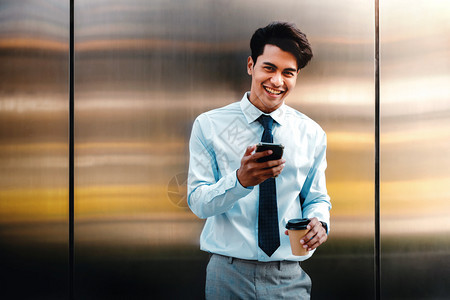 笑交流社会的一位在现代人前线城市生活方式中使用移动电话的快乐青年商人肖像看着摄影机站在长墙边与咖啡杯站立的相片ChoffeCup图片