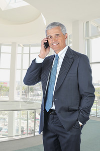 人类中年商站在现代办公大楼里用他的手机进行垂直式交谈在现代化的办公大楼里站立并用他的手机说话成功的经理图片