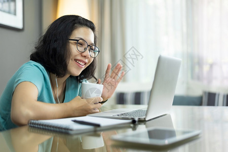 年轻的无线有吸引力亚洲妇女用笔记本电脑和手挥打视频电话身着蓝衬衫的年轻女在家庭网上电子学习和中通过互联网技术与家人和民众交谈穿着图片