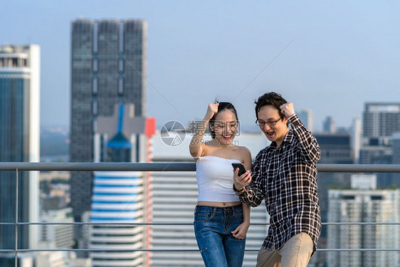 曼谷同业商人和穿便衣妇女正在庆祝他们通过智能手机在商业中心市大楼屋顶上的企业成功实现就概念这是他们共同合作的商人和女随身穿临时西图片