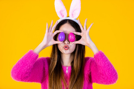 鸡蛋上的眼睛串烧黄色的复活节概念快乐笑亚洲年轻女穿着兔子耳朵的年轻女士在她眼前举着一个多彩的复活节鸡蛋在黄色空版间工作室背景上被孤立宗教背景