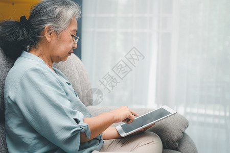 无线的高级亚洲妇女戴眼镜在家沙发上放松并使用平板电脑阅读新闻和子书老年人技术概念为老年人提供技术概念成熟高级的背景图片