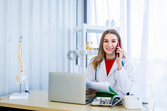 白色的女医生持住拿起智能手机配有听诊器使用笔记本电脑在医院背景的木桌上写文件办公室诊所图片