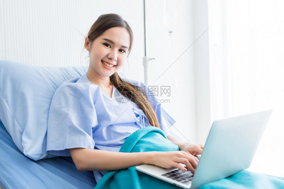 网络亚裔年轻女患者微笑的脸在房间里医院背景的床上用笔记本电脑工作认为在生病时有工作业务诊断亚洲人图片