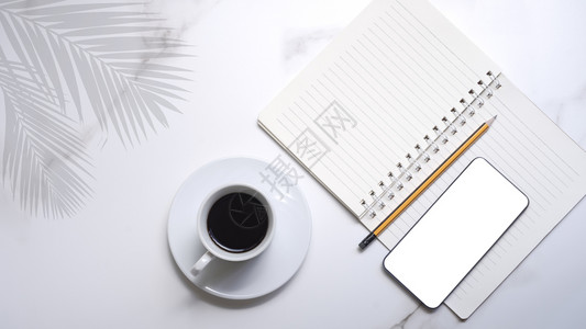 白色陶瓷杯中的黑咖啡智能手机模型带螺旋笔纸的铅和白大理石板上的棕榈叶阴影放松陶瓷制品优质的图片