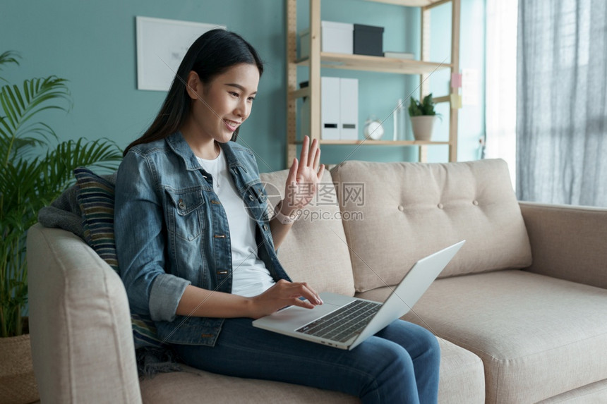 坐着视频活的美丽亚洲年轻女正与同事举行电视会议通过室内技术使用电脑笔记本进行交流在家中使用电脑笔记本图片