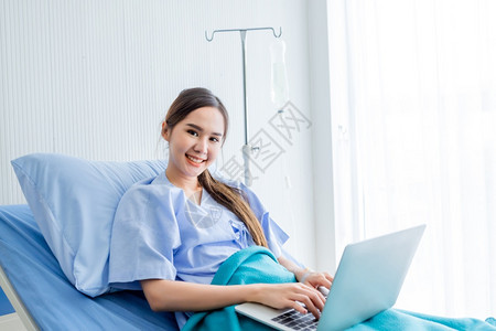 亚洲年轻女病人带着微笑的容在房间里的医院背景中用笔记本电脑在床上工作认为自己生病时有意可做恢复吸引人的药物图片