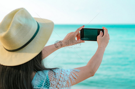 年轻亚洲妇女戴帽子时使用智能手机在热带海滩的沙上拍照在热带天堂海滩暑假在热带天堂海滩举行夏日度假快乐的嬉马女孩在节假日旅行时享受图片