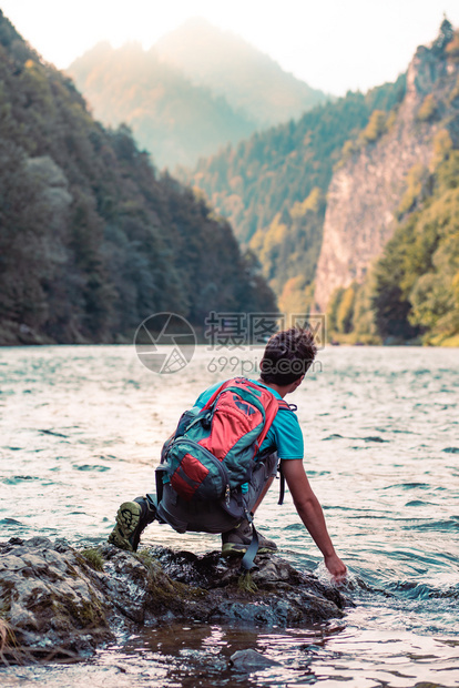 背着包坐在河边岩石上手握在水中看着山峰的年轻男孩他穿着运动夏季服装户外降低抓住图片