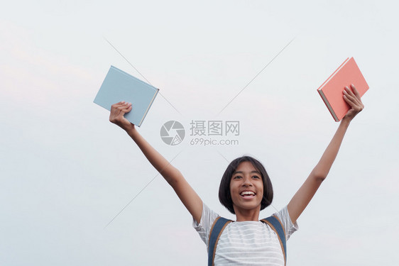 快乐的亚洲女孩在拿着一本书和手举到蓝天背景时脸上微笑容瞳孔学习生图片