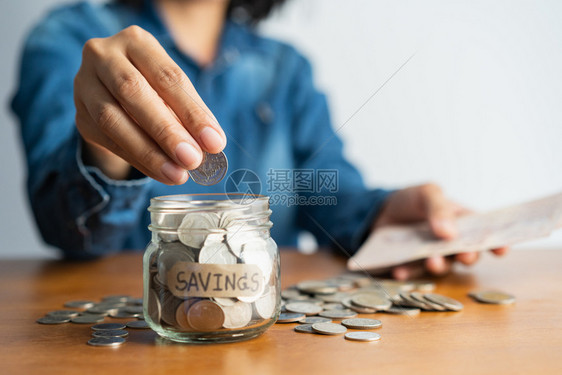 贷款女人手在玻璃瓶里放一枚硬币在棕色木制桌子上放一堆硬币投资企业退休金融为未来概念存钱铢收入图片