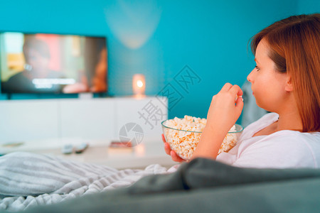 白种人坐在家里睡沙发床上的年轻妇女躺在一个爆米花碗上躺着在她单独享受电影或系列的公寓里看视用毯子覆盖电视美丽的食物图片