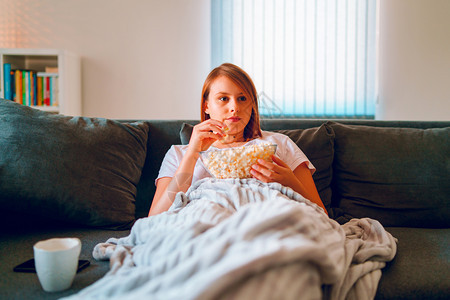 坐在家里睡沙发床上的年轻妇女躺在一个爆米花碗上躺着在她单独享受电影或系列的公寓里看视用毯子覆盖电视娱乐放松长椅图片