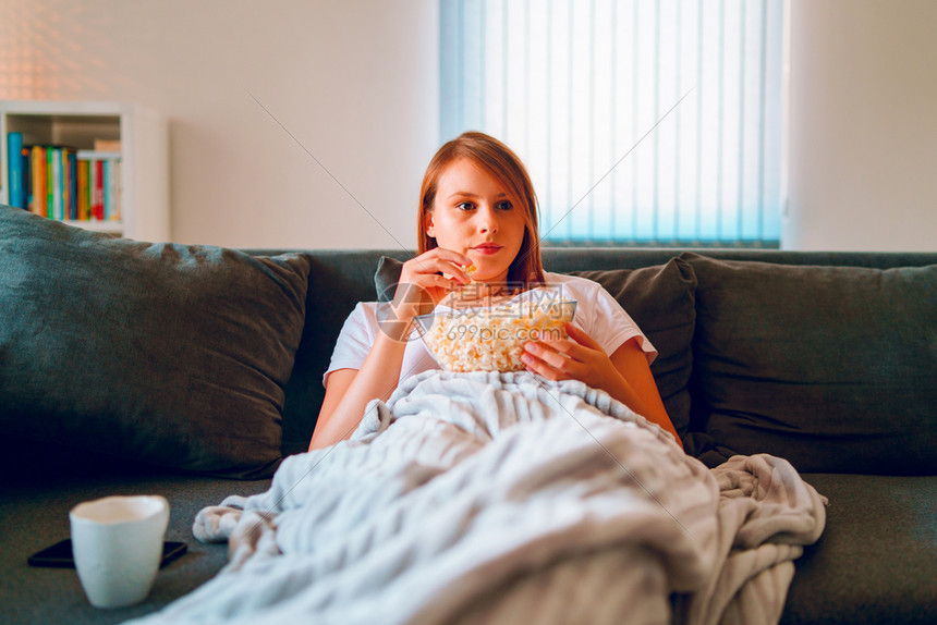 坐在家里睡沙发床上的年轻妇女躺在一个爆米花碗上躺着在她单独享受电影或系列的公寓里看视用毯子覆盖电视娱乐放松长椅图片