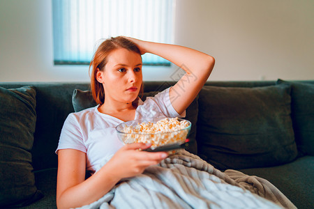喜剧保持视频坐在家里睡沙发床上的年轻妇女躺在一个爆米花碗上躺着在她单独享受电影或系列的公寓里看视用毯子覆盖电视图片