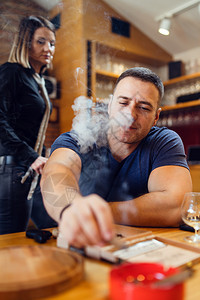 随意的雪茄餐厅坐在桌边着的年轻天主教男子肖像在餐馆或家中穿着蓝色T恤衫看着烟灰盘而冒的无名女子走路或站在他身后时扑灭了在餐馆或家图片
