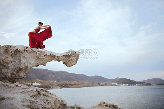 舞蹈家身穿红礼服的黑发年轻女人在海滨附近岩石上跳舞海浪靠近图片